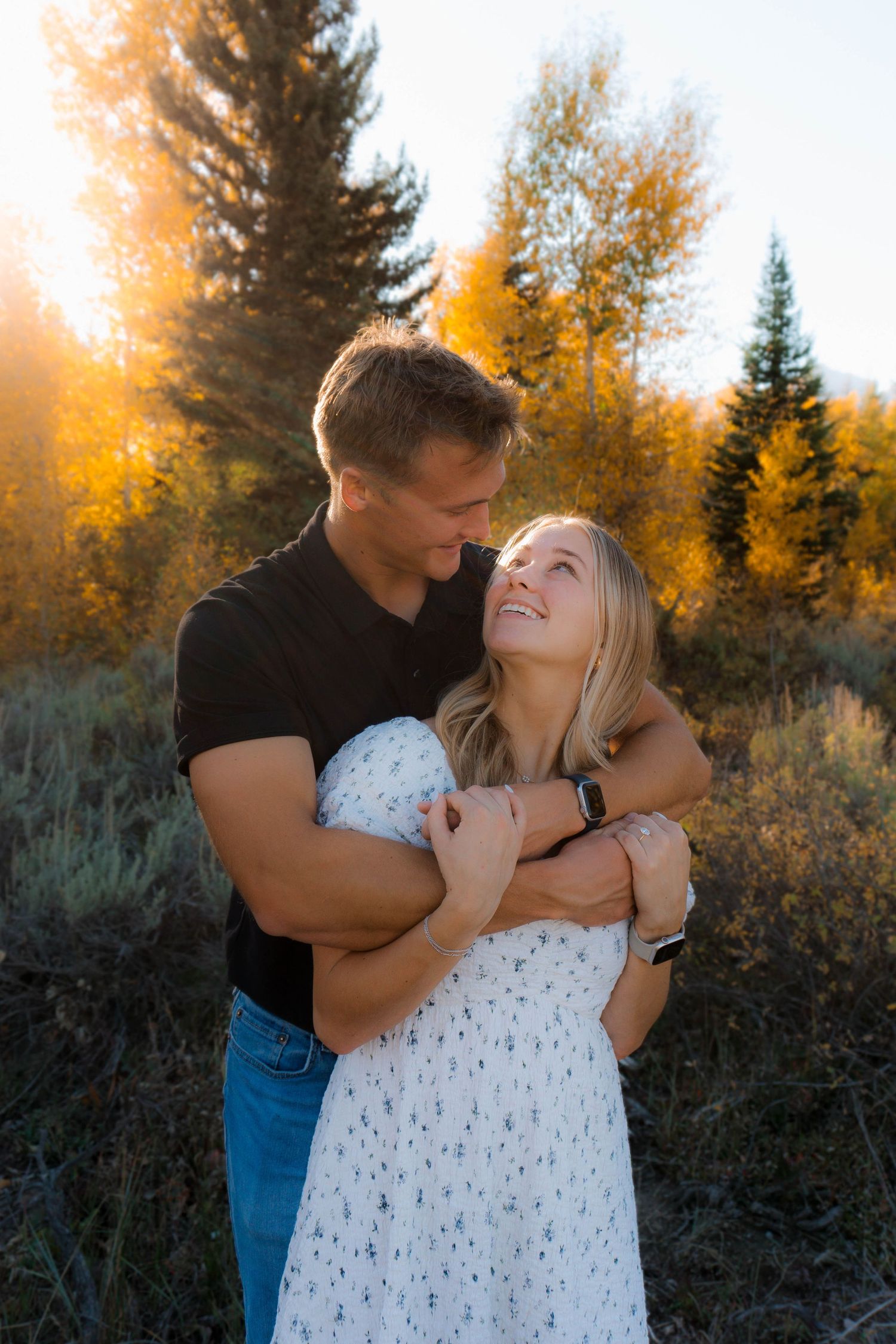 Romantic couple embraces against golden autumn sunset with evergreen trees in background.