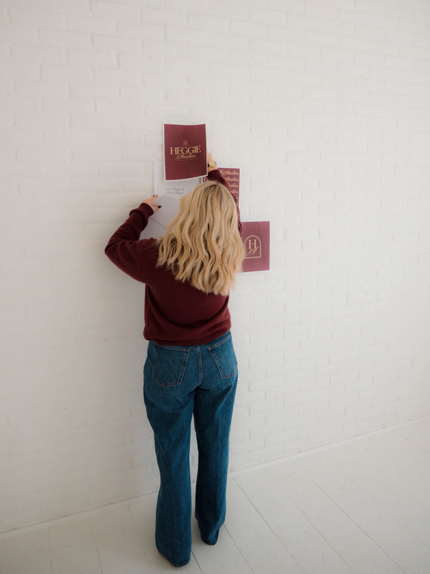 Person wearing burgundy sweater and jeans leans against white wall while reading small red book.