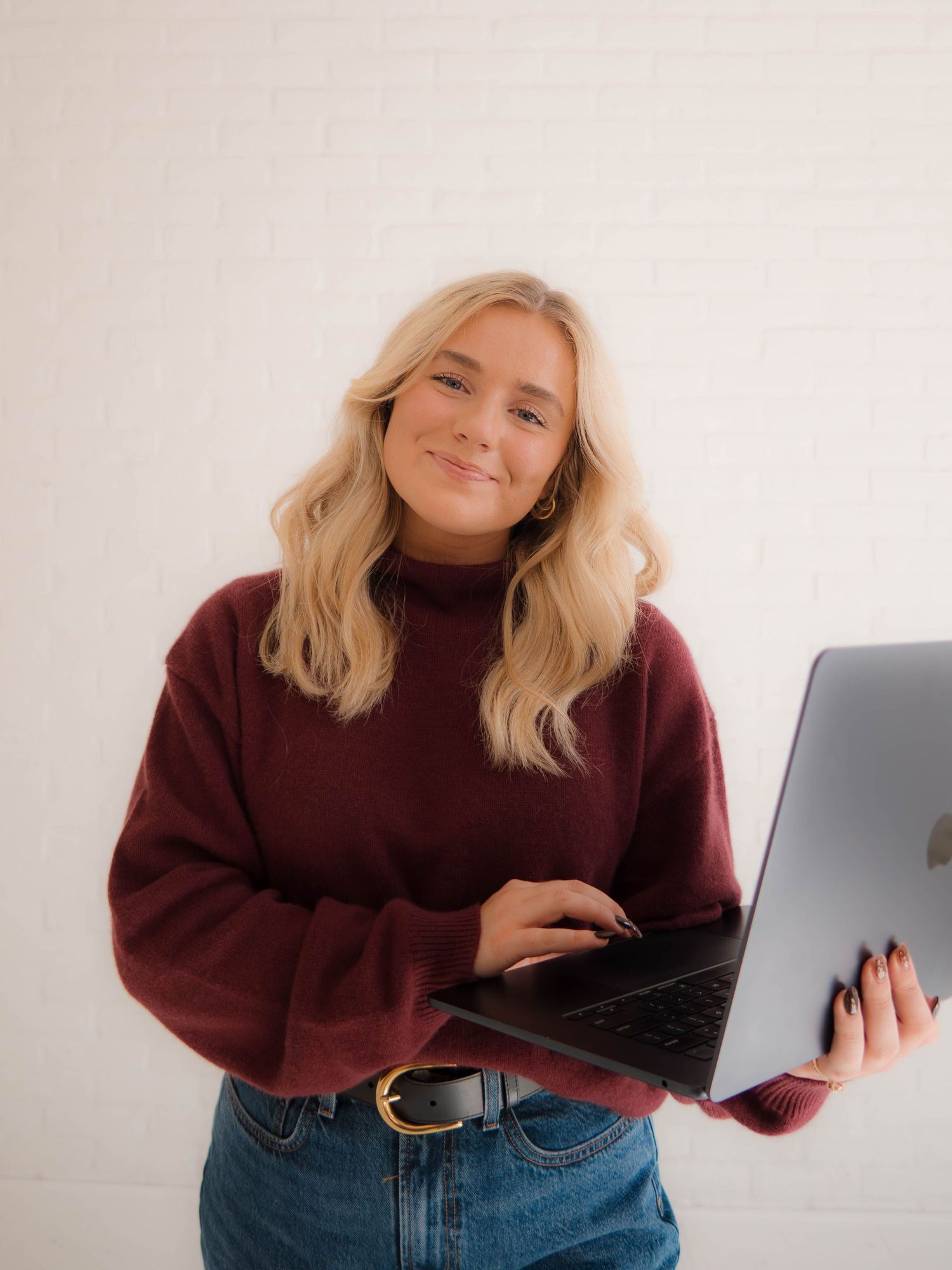 Person in burgundy sweater and jeans works on laptop while sitting on floor against white wall.