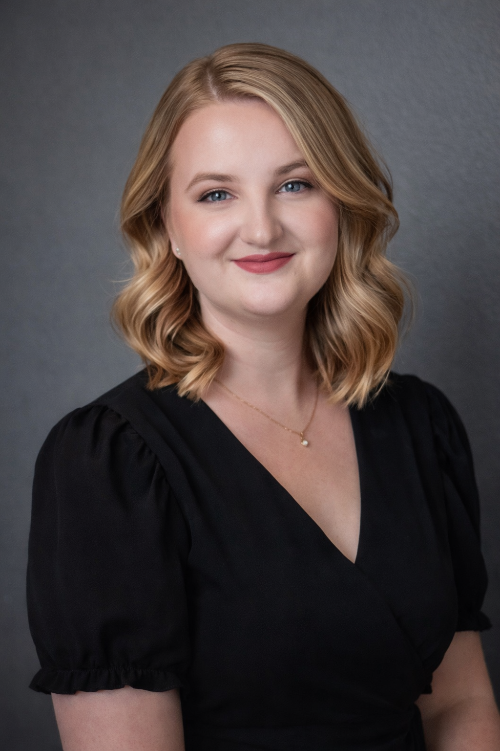 Professional headshot portrait of a person in a black v-neck dress with puff sleeves against a gray background.
