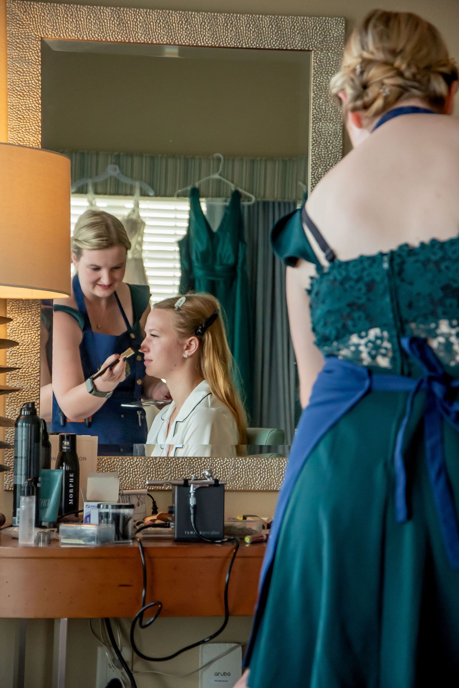 A makeup artist applies cosmetics at a vanity while someone in a teal formal dress stands nearby.