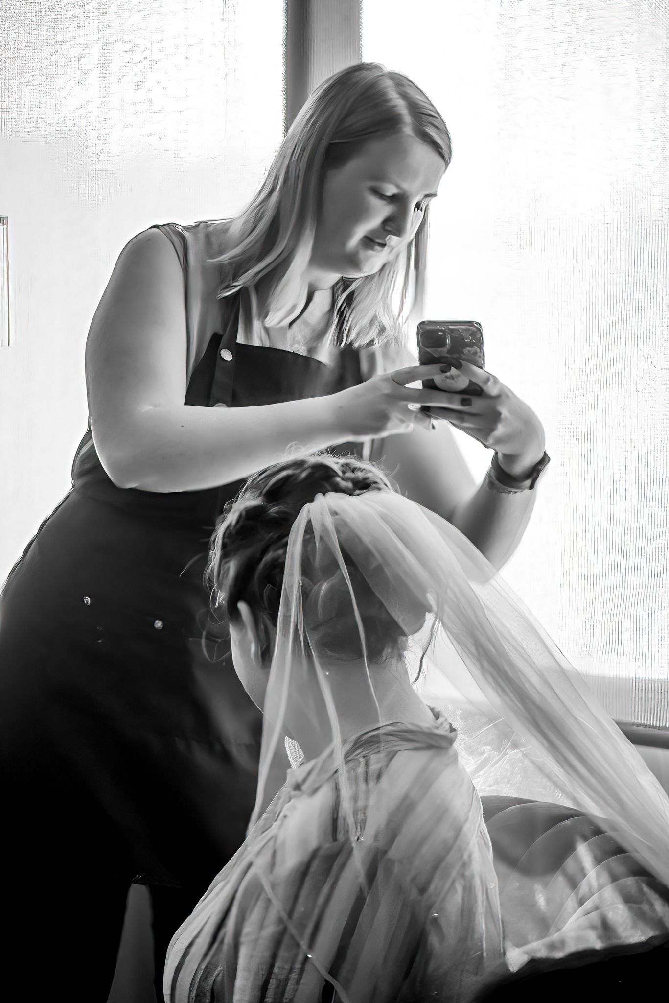 Black and white photo in an Ocala, FL salon of a blonde woman taking a photo with her phone of a bride with her hair done, wearing a veil.