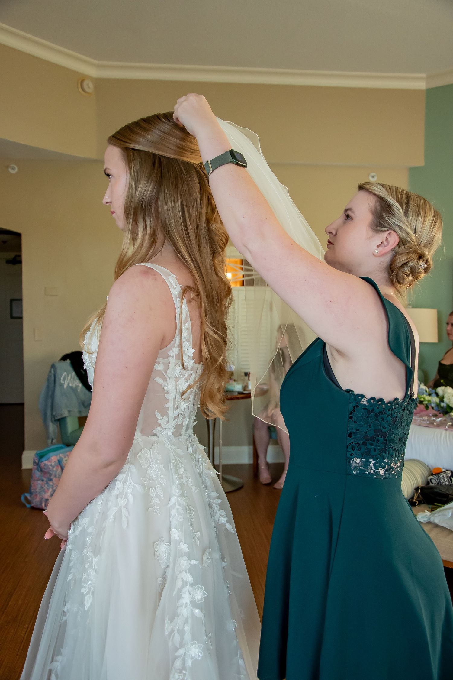 A person in a teal dress helps adjust the veil on a bride wearing a white lace wedding gown during preparations.