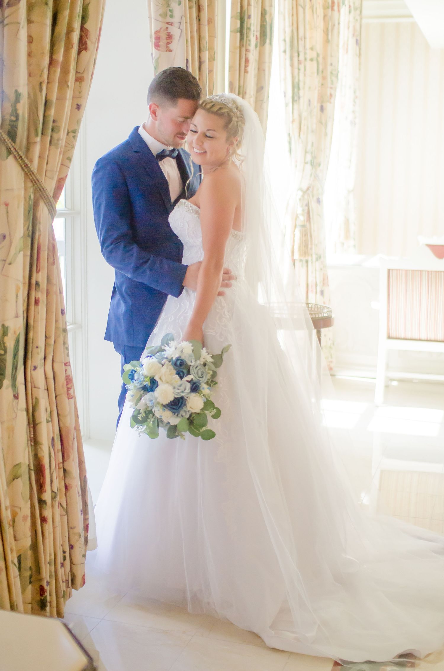 A bride and groom in a loving embrace. Navy blue suit, white wedding dress, veil blue and white floral bouquet. Copyright Vazul Studios.