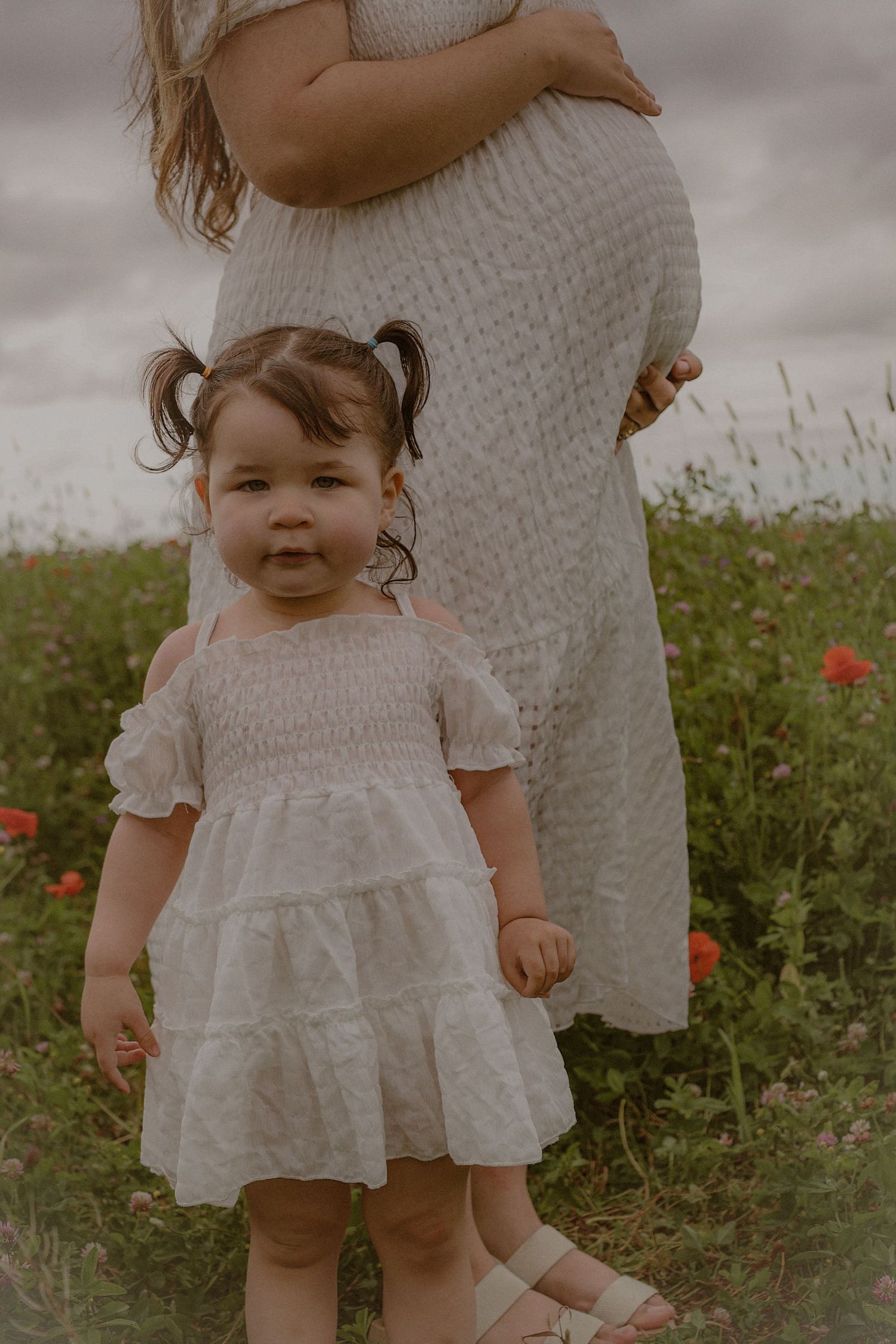 Maternity photography outdoors in a poppy field, featuring a mother and her toddler daughter and bump, Moody skies create cinematic feel