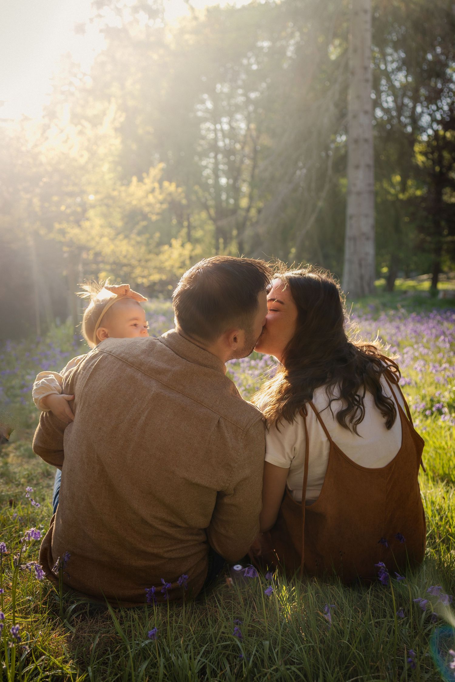 Portrait of parents sharing a kiss, holding newborn daughters and their toddler daughters during a spring, evening in a bluebells field