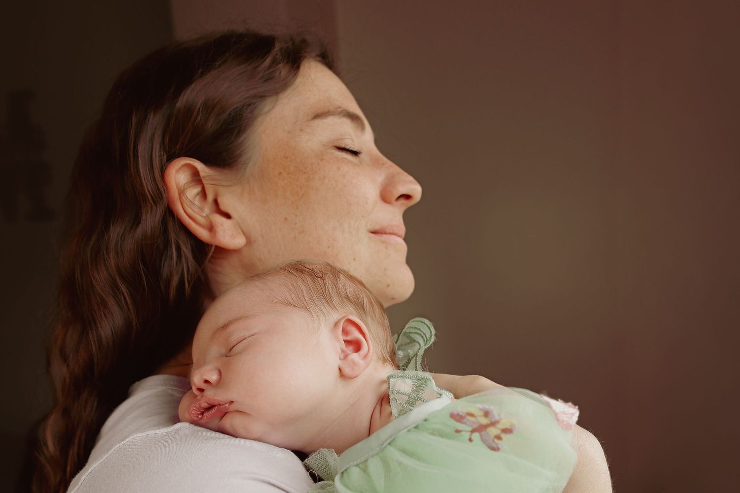 Emotive portrait of mother and newborn daughter sharing a moment of peace, baby sleeps resting on mothers shoulder, mum closes eyes smiling