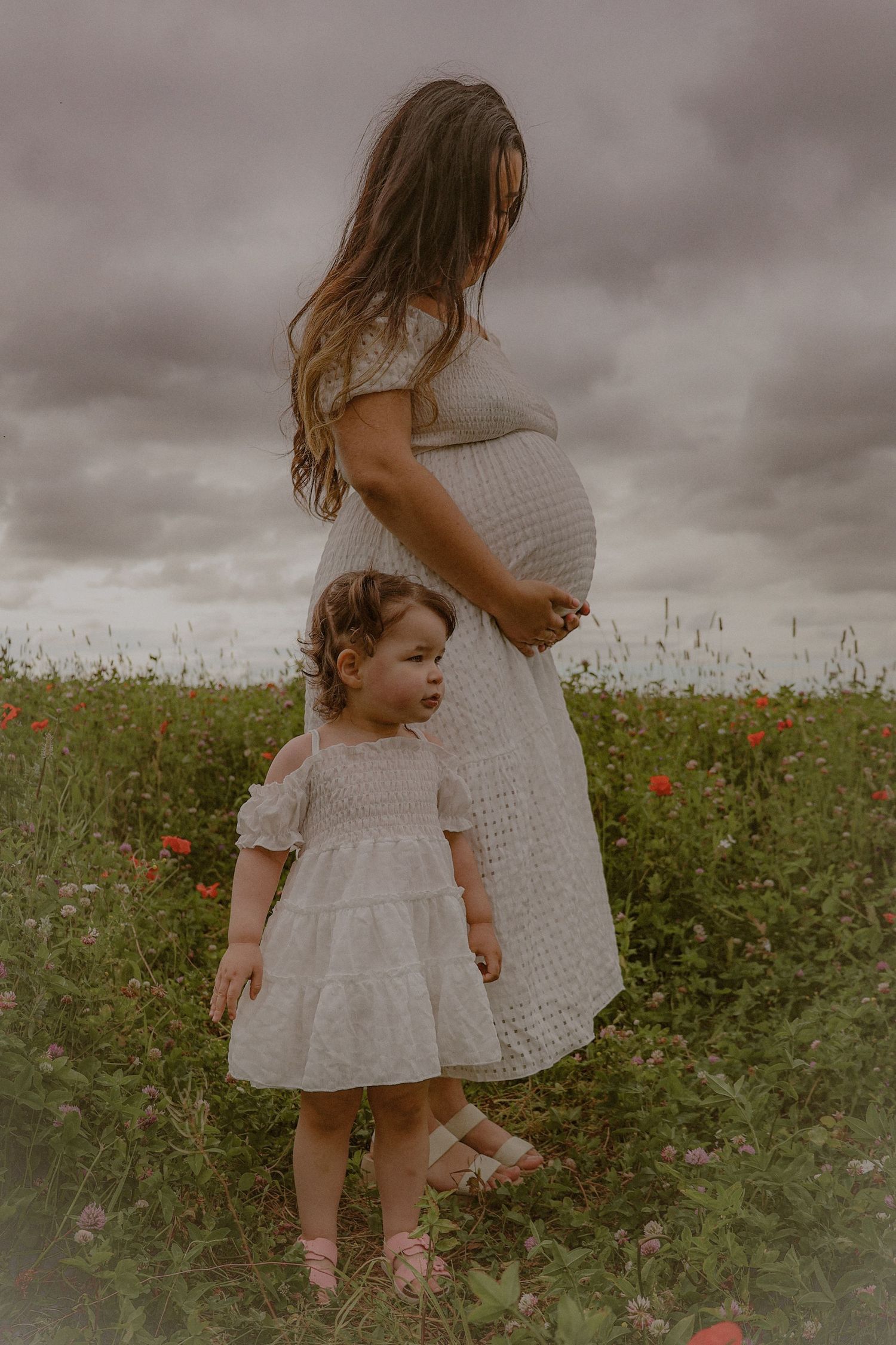 Maternity photography outdoors in a poppy field, featuring a mother and her toddler daughter and bump, Moody skies create cinematic feel