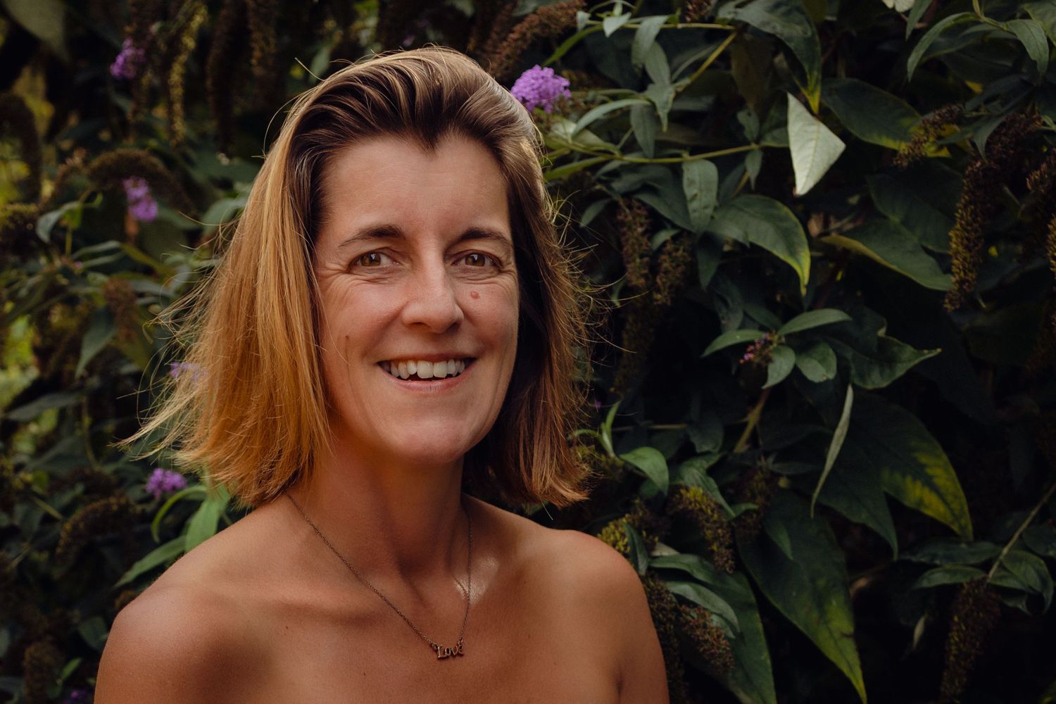 gorgeous woman in nature ,bare shoulders, flowers behind her, shes smiling and buzzing with quiet confidence and inner peace, colour, natura