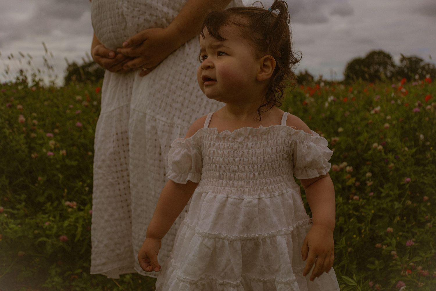 cinematic portrait close up of little girl with mums hands holding her pregnant belly in the background alongside wild flowers, in Norfolk