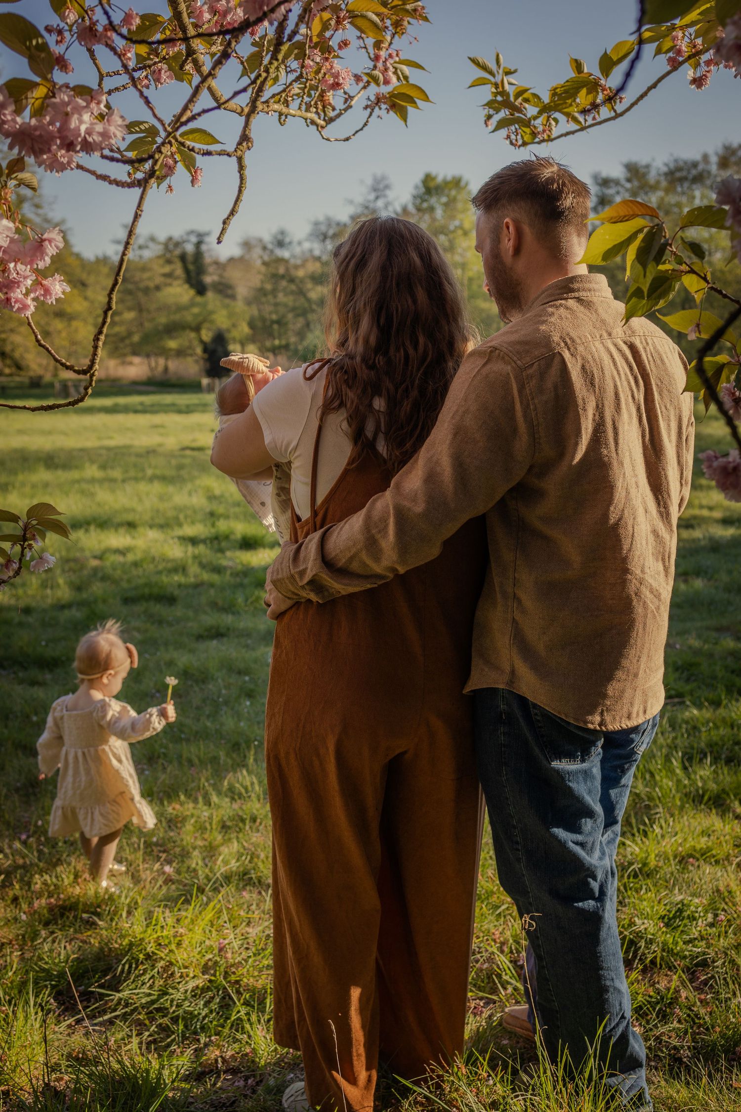 evening outdoors newborn photoshoot, parents watching toddler playing int he field while holding newborn daughter, flowers surrounding them