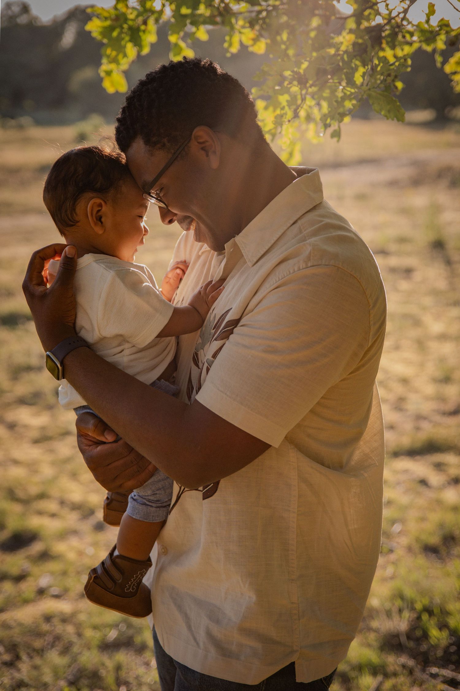 Golden hour portrait of father and baby son by a tree, their foreheads together sharing a candid moment captured by Untamed Muses