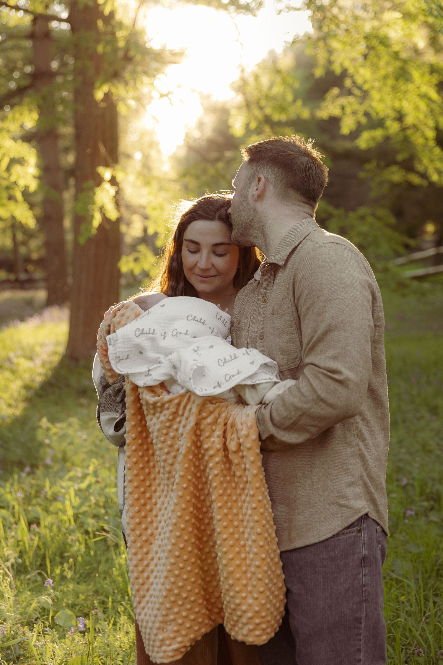 Evening, spring outdoors natural newborn photoshoot, portrait of parents holding baby daughter gazingly lovingly upon her, dad kissing mum