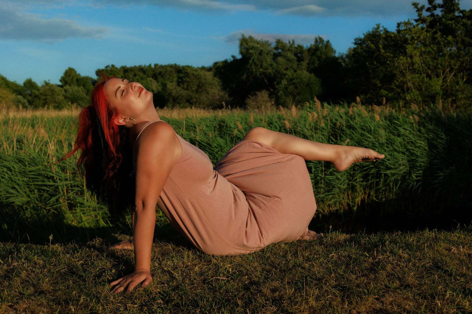 portrait of woman in a field, relaxing with sun in her face ,wind sweeping her har, wearing a dusky pink dress , in Norfolk riverside