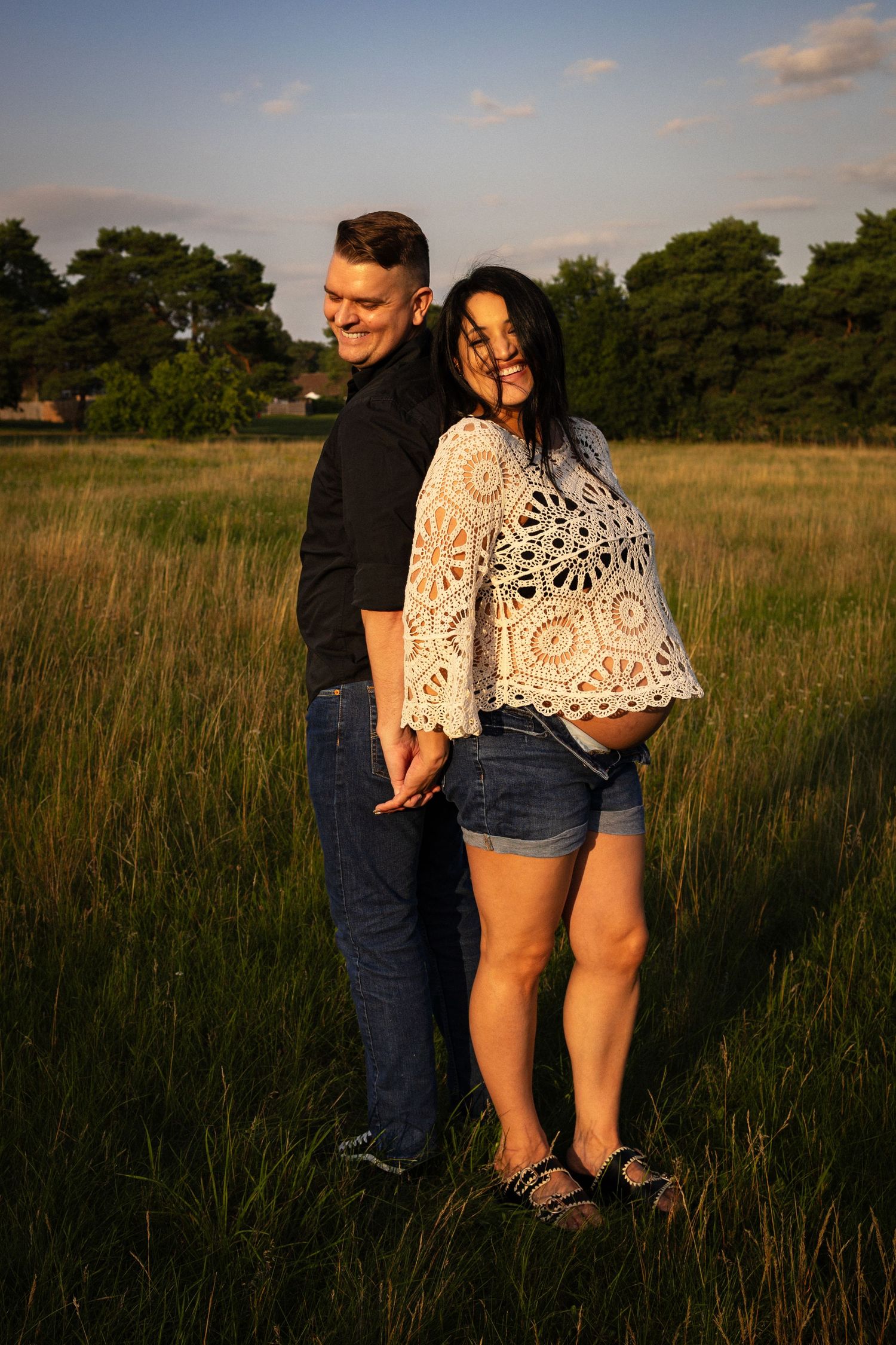 Portrait of joyful expecting parents holding hands back to back in a. Norfolk field  during a summer maternity photoshoot by Untamed Muses