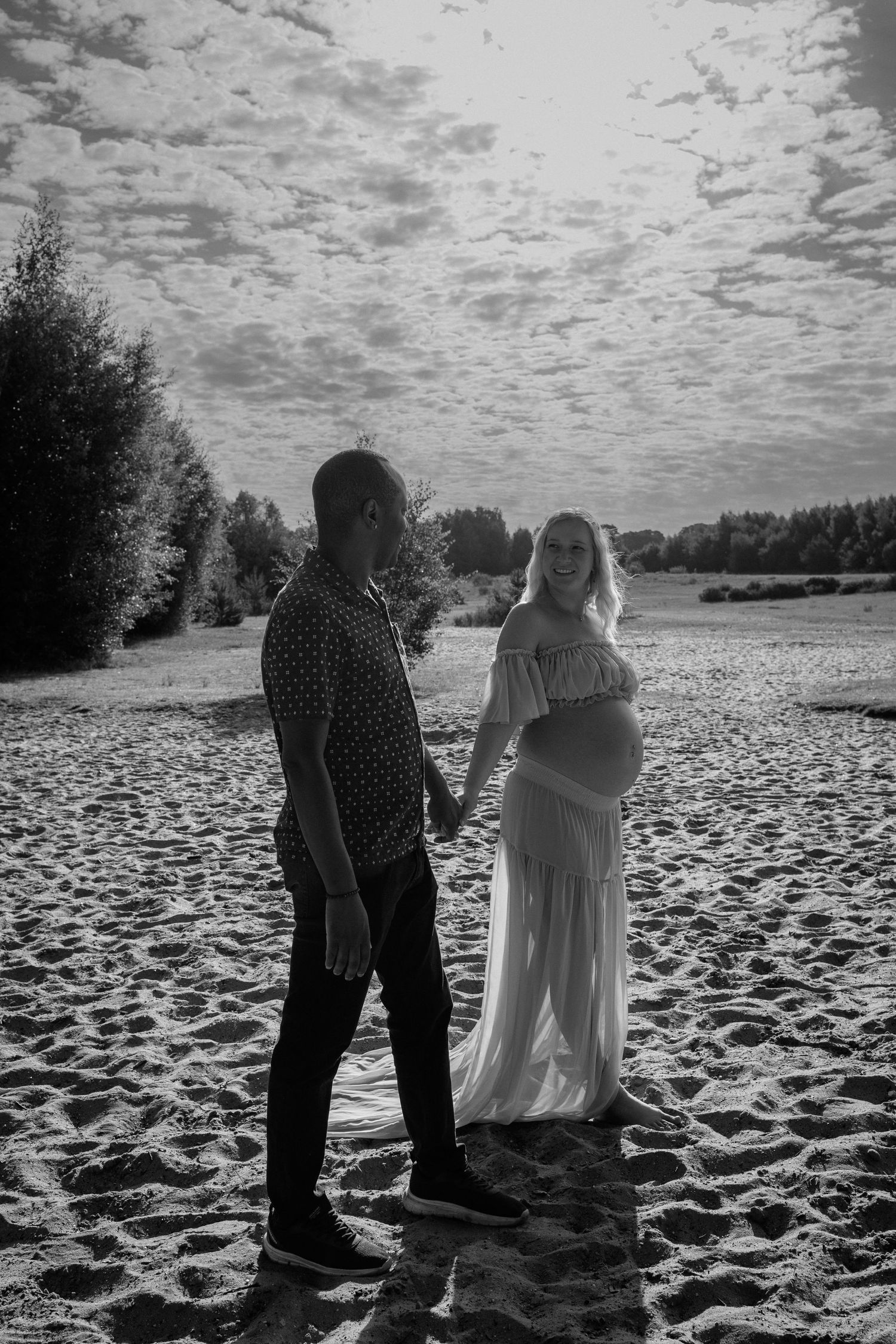 Monochrome portrait of couple holding hands, starting into each other as the walk through a lake setting during maternity photoshoot