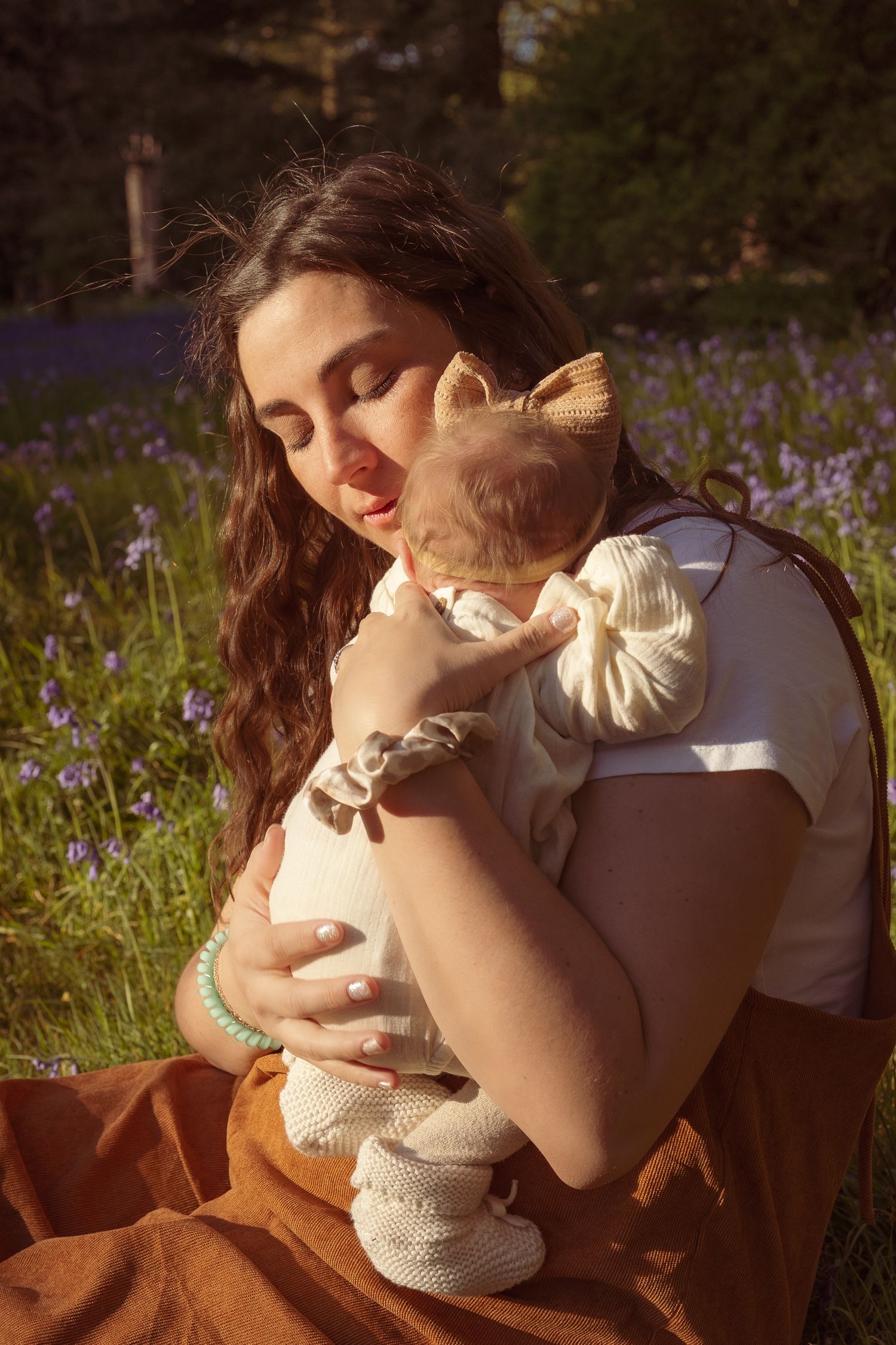 Mother gentle holding newborn daughter in a bluebell field, during golden hour in Norfolk
