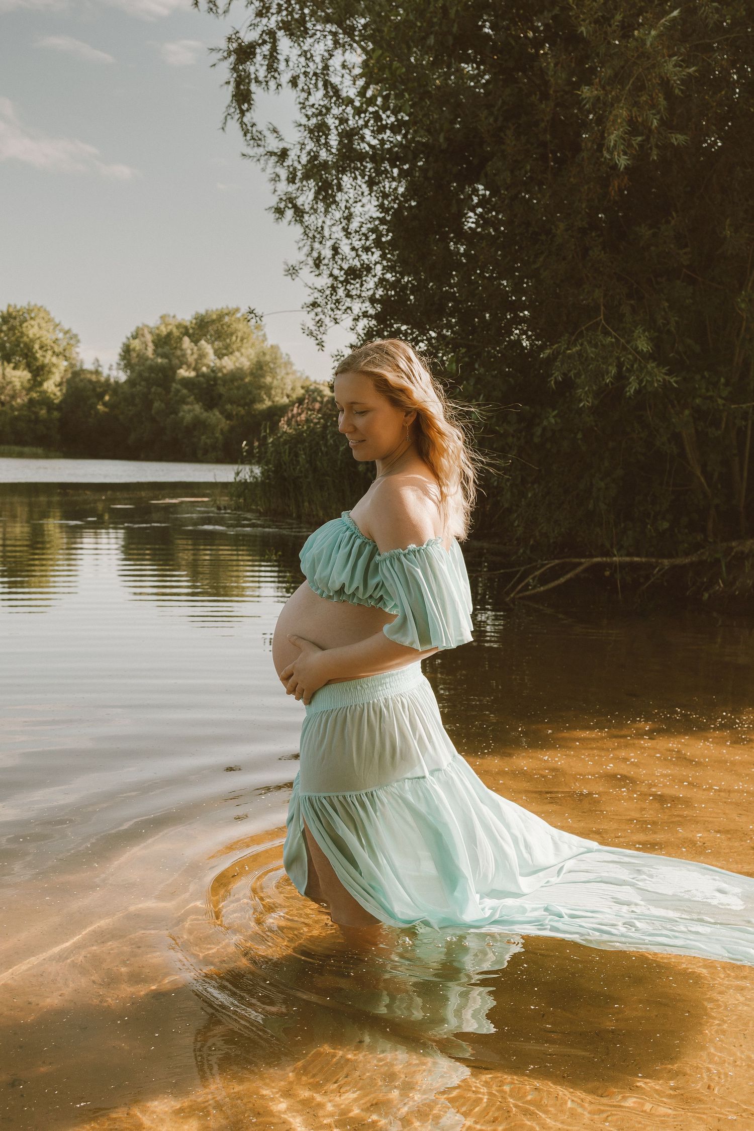 Portrait of beautiful pregnant woman in a lake, wearing a light blue dress, looking down at her bump boringly by empowerment photographer