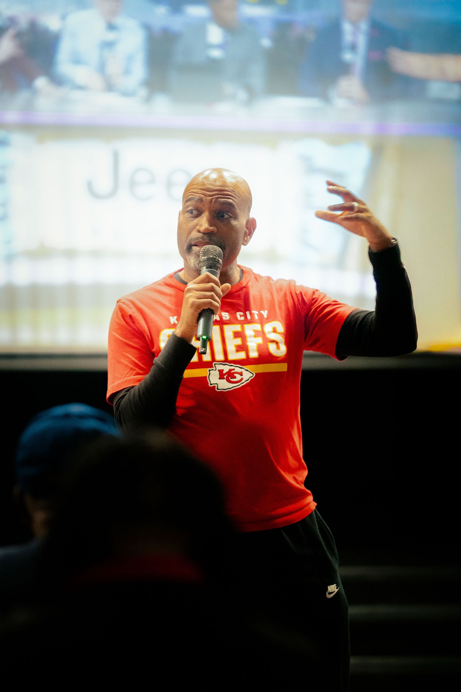 A speaker in a Kansas City Chiefs shirt energetically addresses an audience during a presentation.