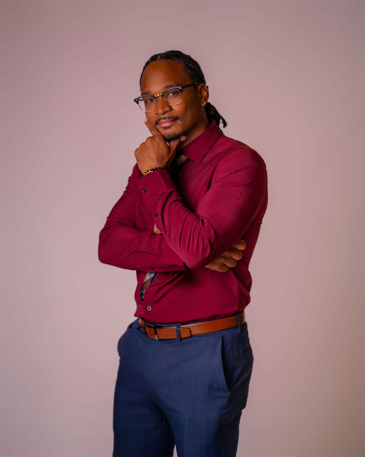 Professional man in burgundy shirt and blue pants posing confidently with arms crossed against neutral background.