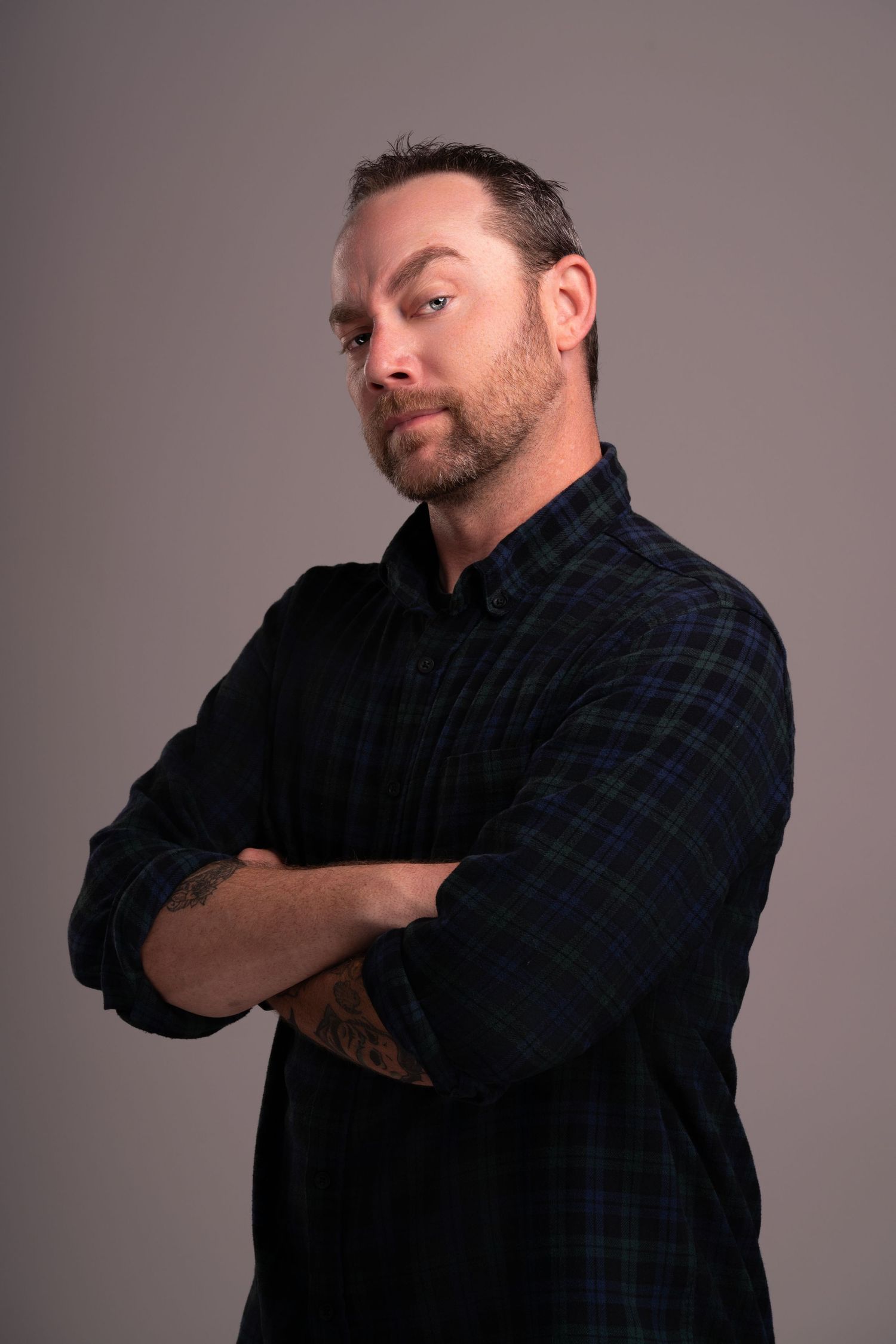Side-angled portrait of person in plaid shirt with crossed arms and facial hair.