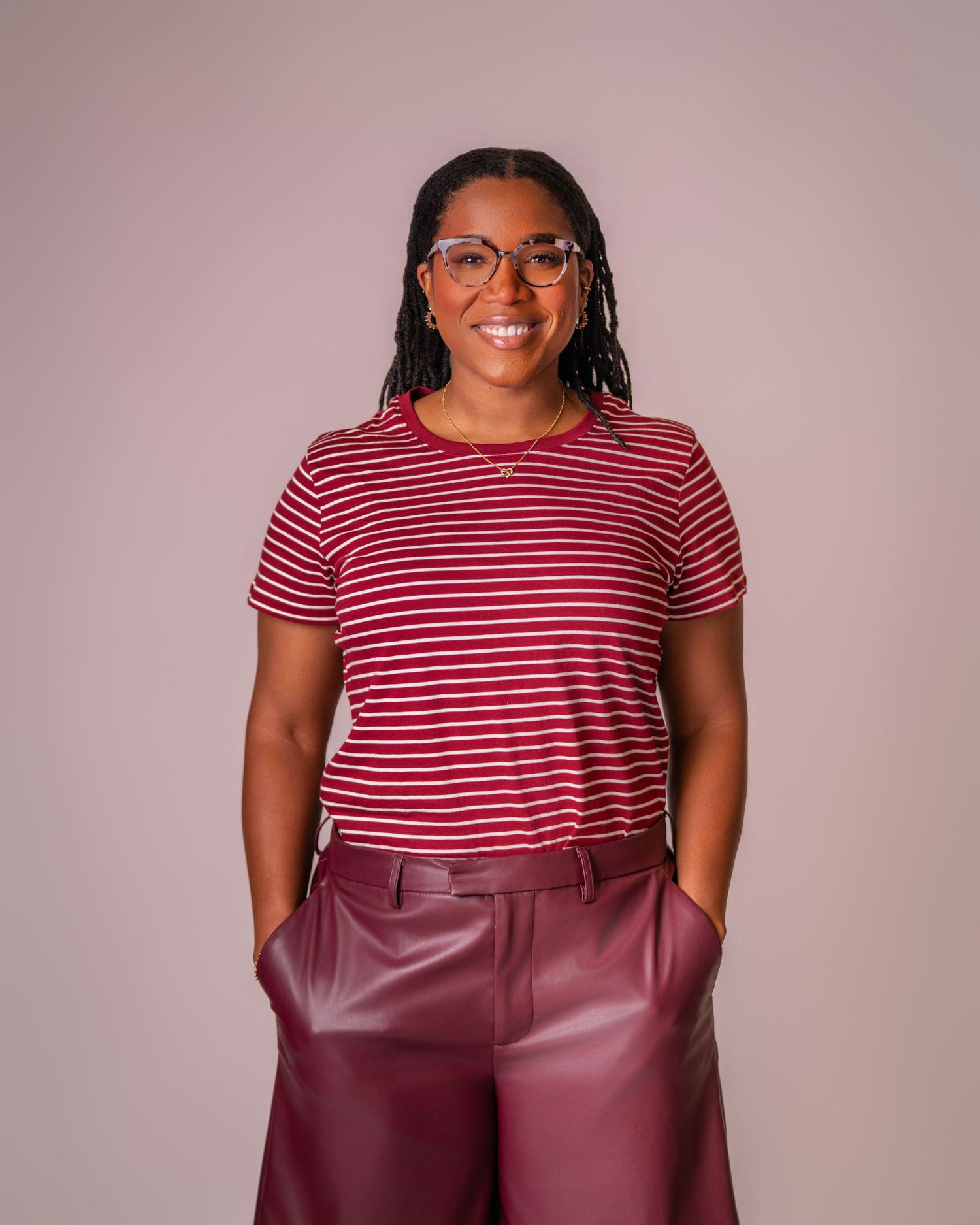 Smiling woman wearing red striped t-shirt and burgundy leather pants, posing with hands in pockets against pink background.