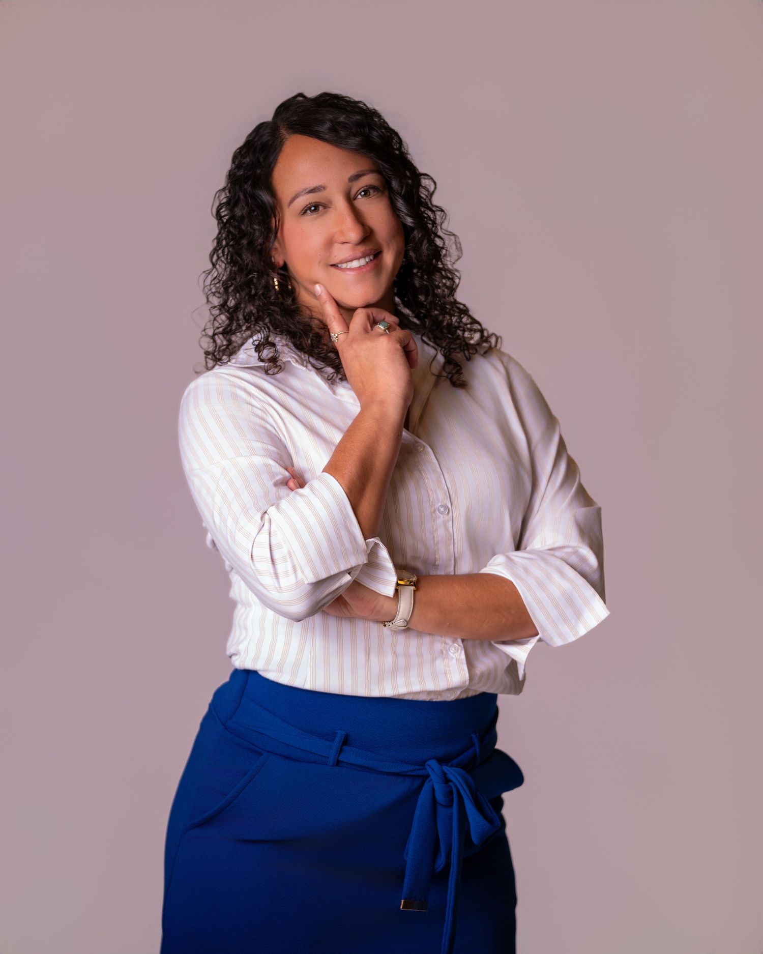 A confident woman with curly hair poses in a white striped shirt and blue wrap skirt against a neutral background.