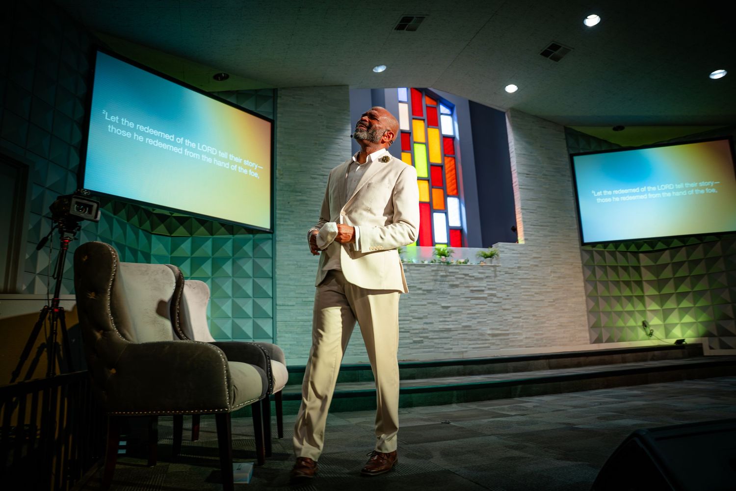 A speaker stands on stage in a beige suit delivering a presentation with projection screens and colorful wall decor visible.