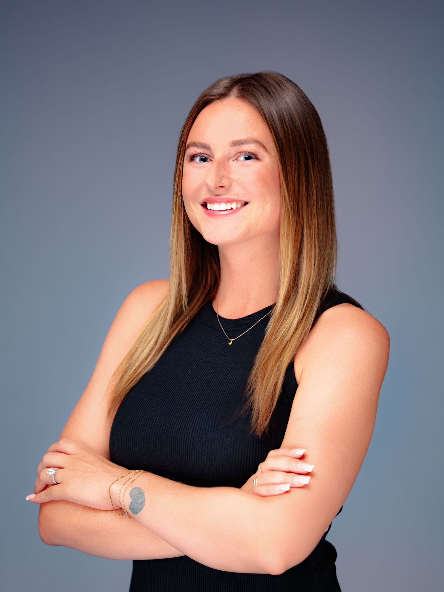 Professional portrait of a confident businesswoman in a black sleeveless top with long ombre hair against a gray background.