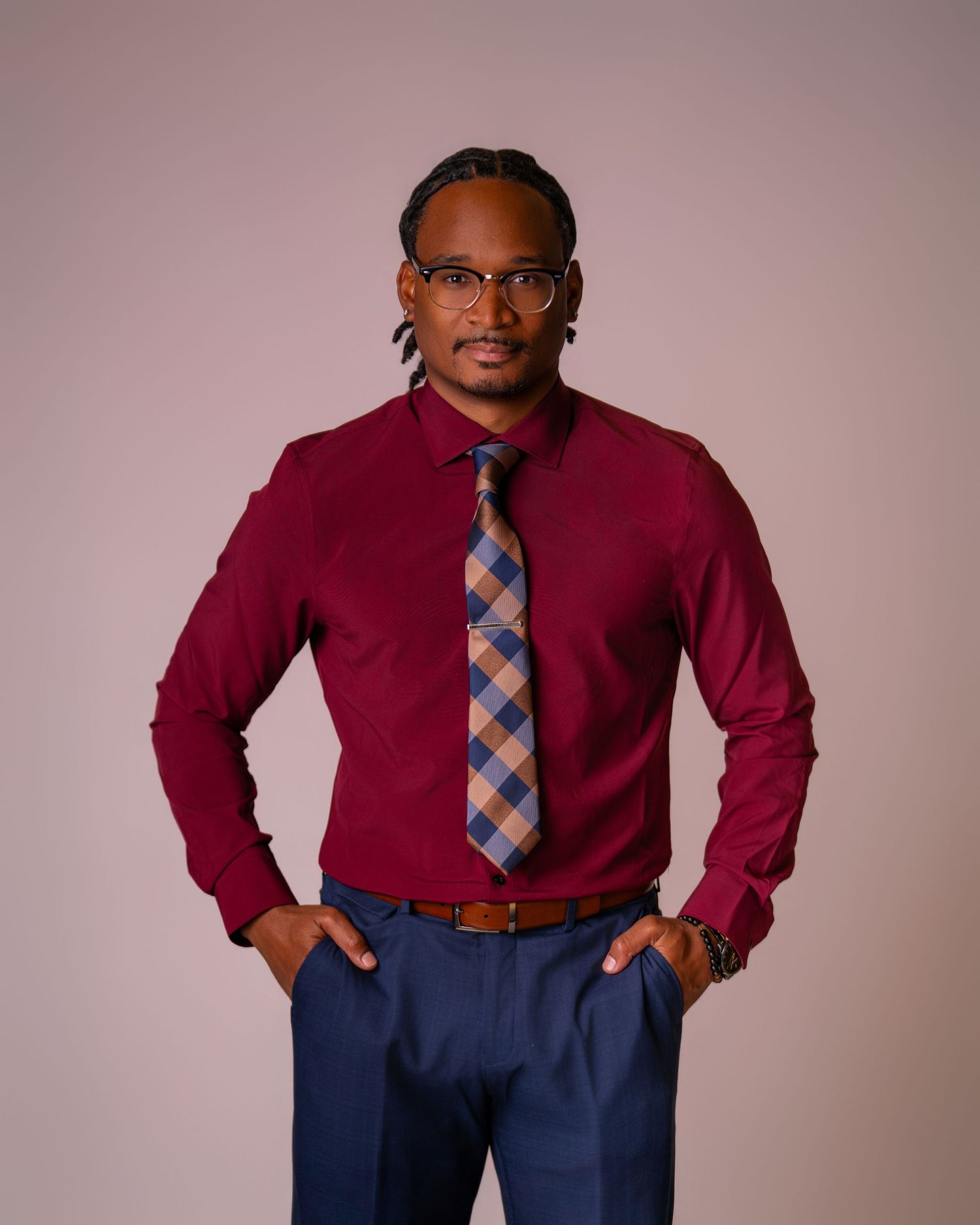 Man in burgundy dress shirt, plaid tie, and navy blue pants poses confidently with hands in pockets.