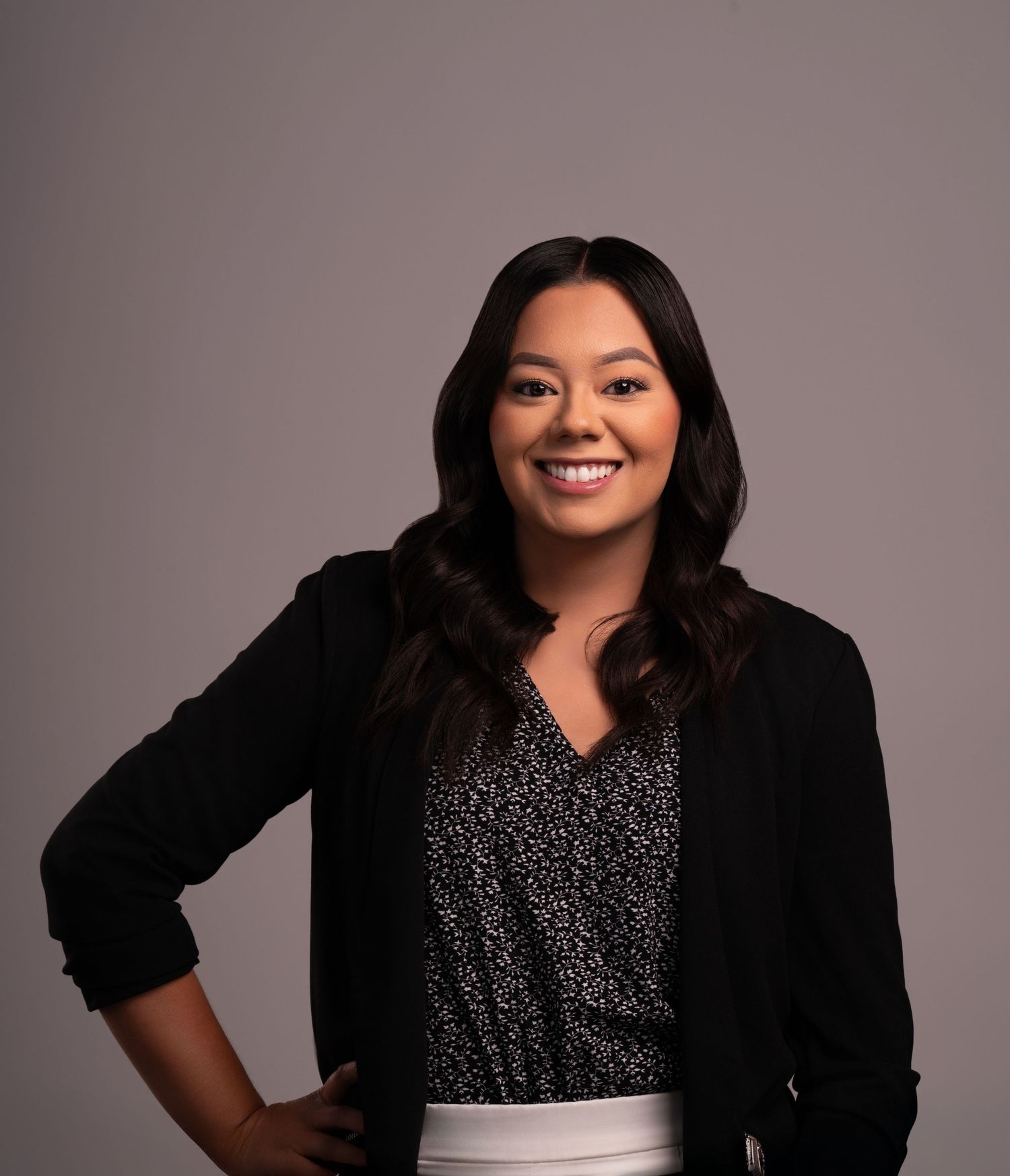 Professional headshot of someone wearing a black blazer and patterned top against a gray background.