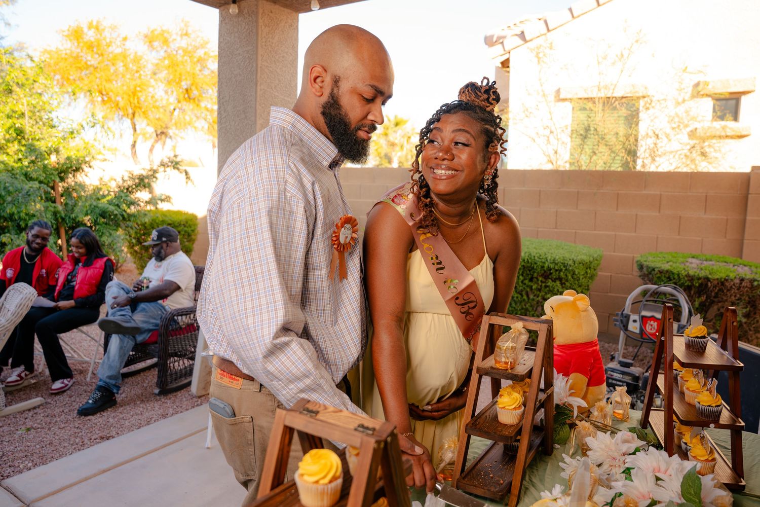 Couple sharing a joyful moment at an outdoor celebration, woman wearing a sash, guests seated in background.