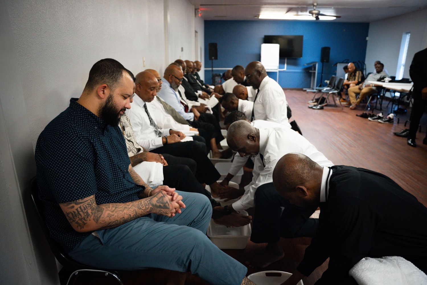 A solemn prayer gathering shows people in reverent poses seated in chairs.