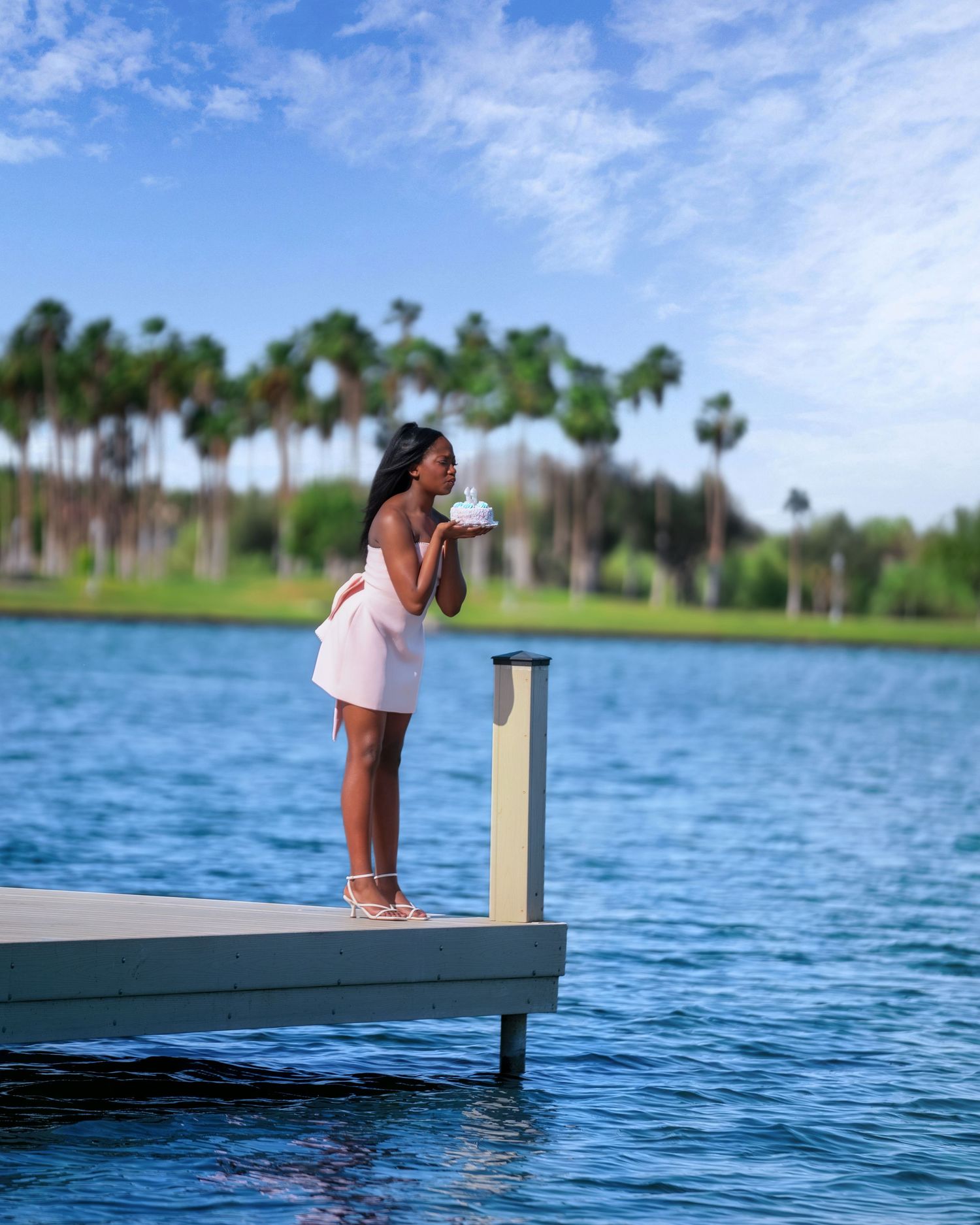 A woman in a white dress stands on a dock blowing a kiss, with palm trees and blue water in the background.