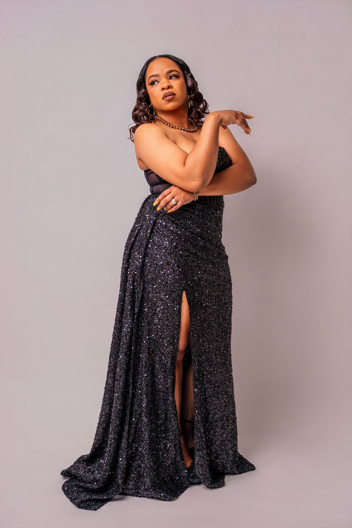 Model in sparkling black evening gown poses elegantly against neutral backdrop in fashion photoshoot series.