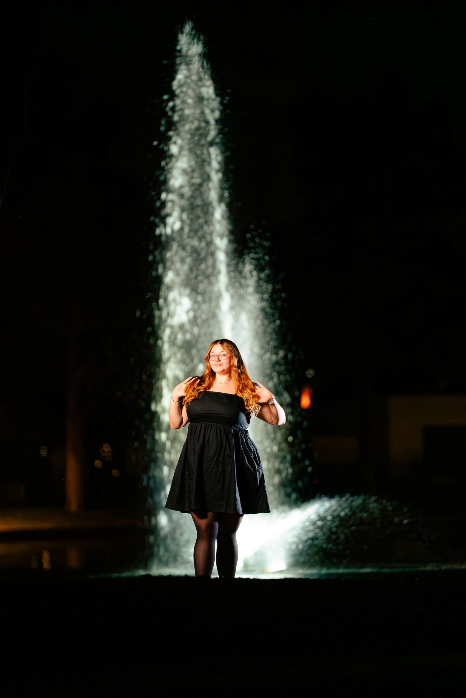 Woman in black dress stands before illuminated fountain at night, red hair flowing, elegant outdoor portrait.