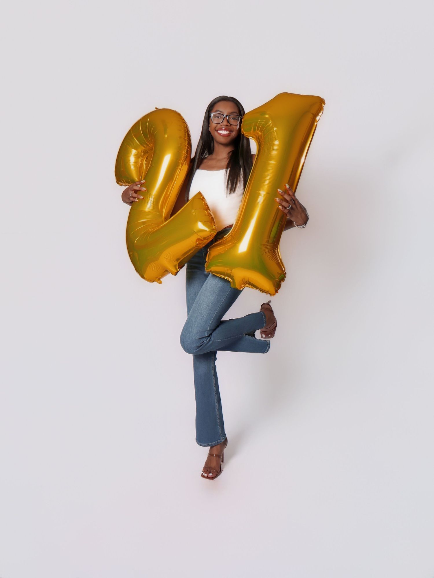 Celebratory pose with large gold number 21 balloons while wearing jeans and white top.