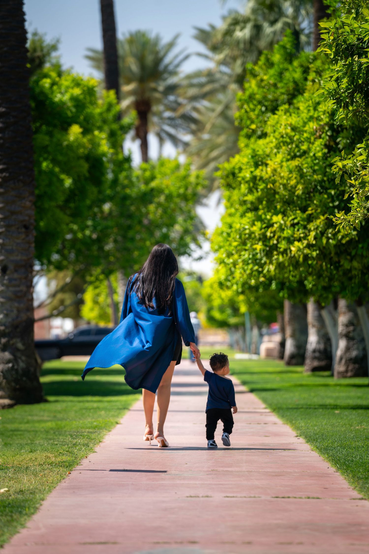 Person in blue dress walks with small child along palm tree-lined pathway.