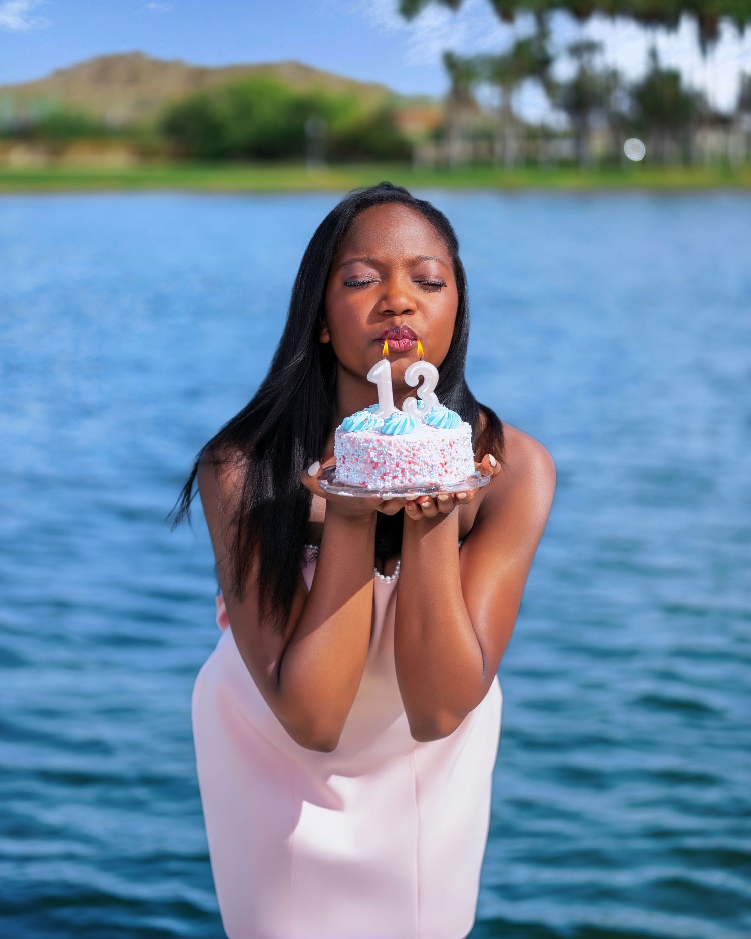 Woman in pink dress blowing candles on a small birthday cake with number 12, standing by a scenic lake.