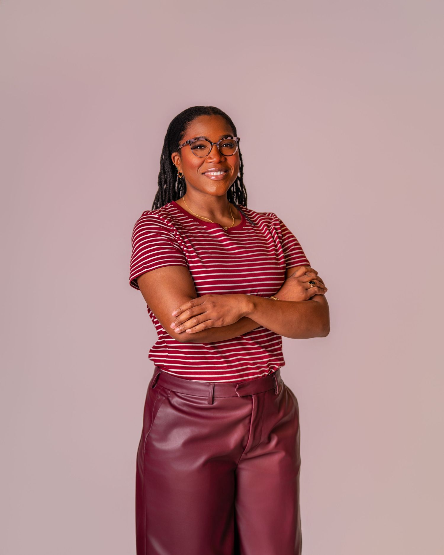 Woman in striped red top and burgundy leather pants poses confidently with arms crossed against pink background.