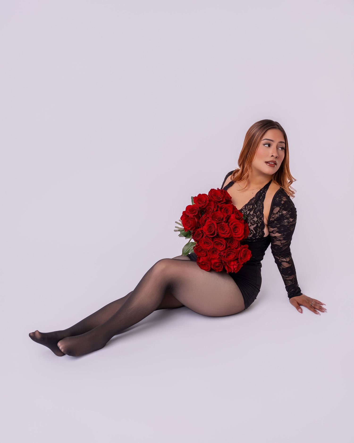 A woman in black lace bodysuit poses with red roses against a light gray backdrop.