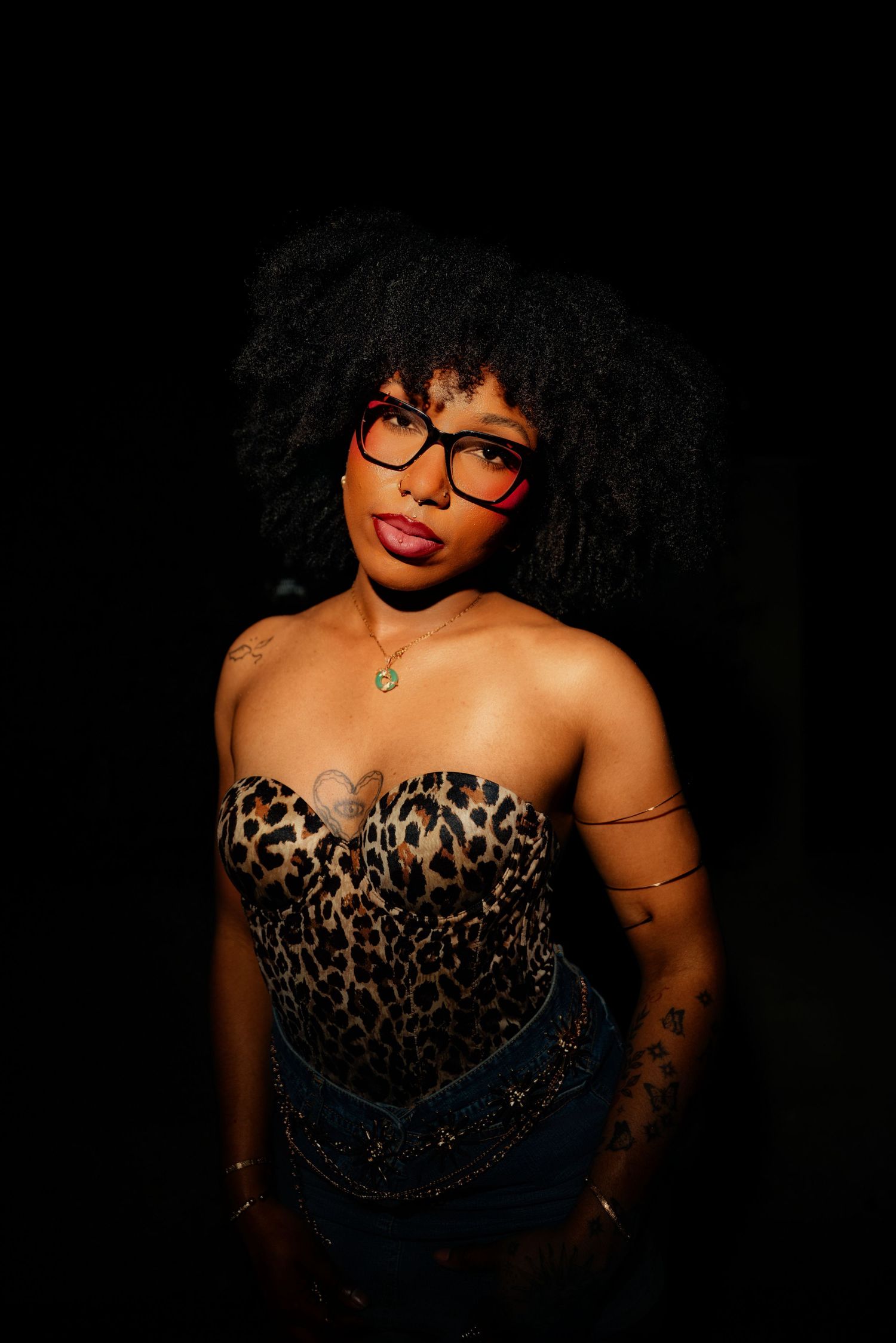 Woman with afro hair wearing red glasses and leopard print corset poses confidently against dark background.