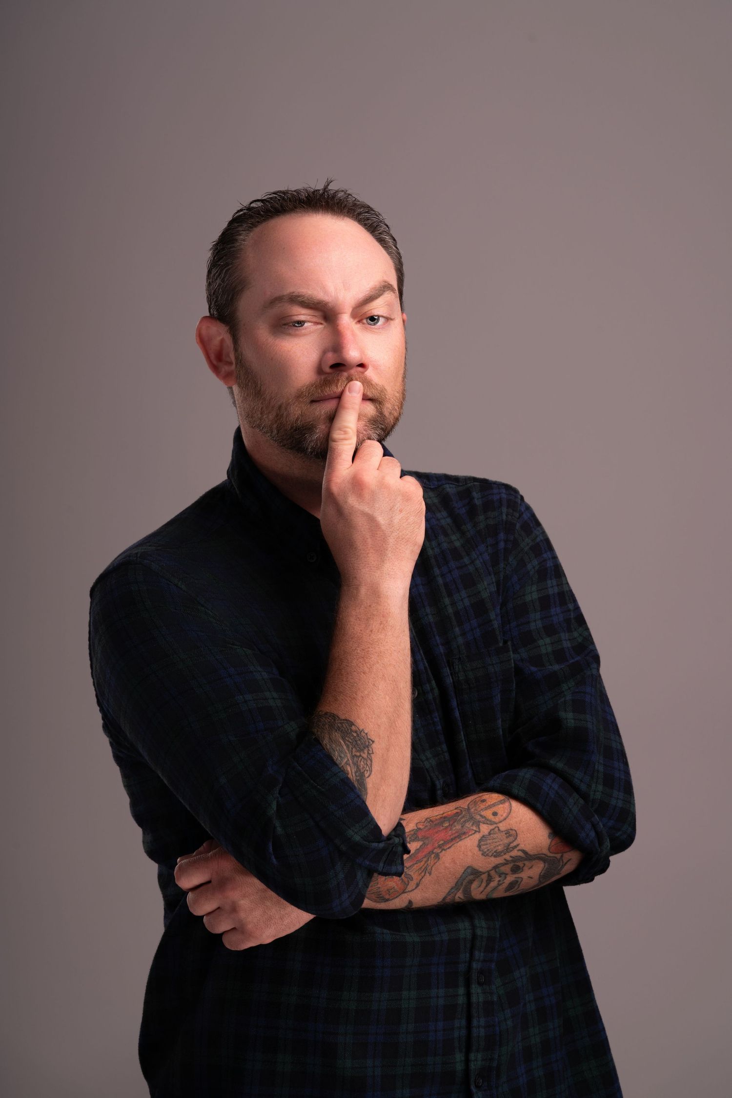 Thoughtful pose in dark plaid shirt with hand near face showing visible arm tattoos.