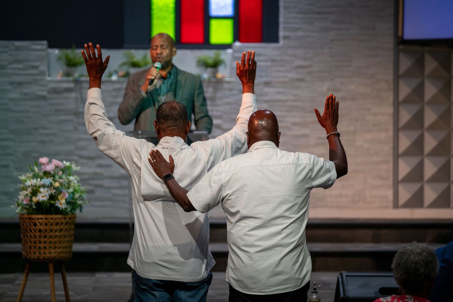 People worship with raised hands during a church service with colorful stained glass windows visible.