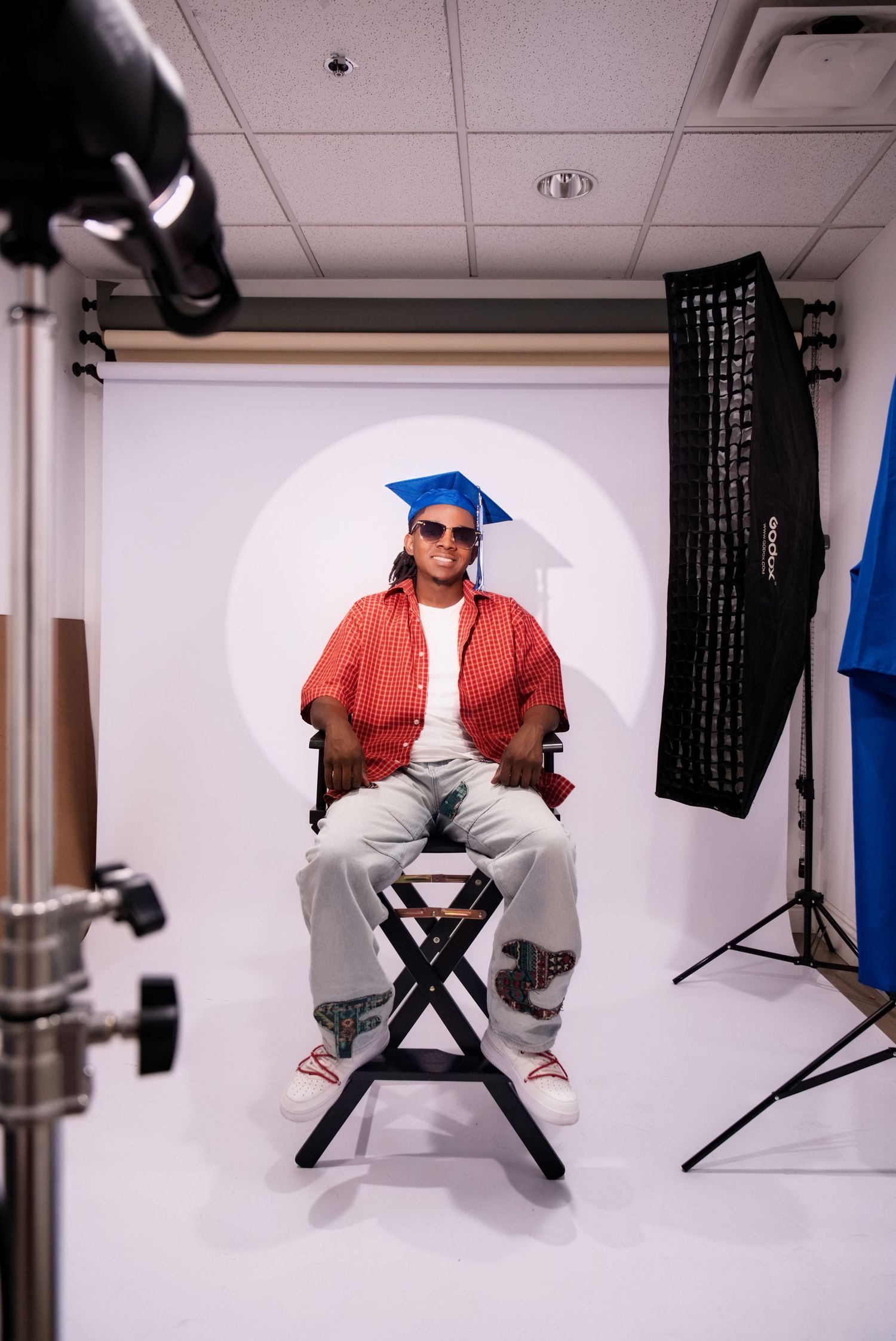 Graduate in blue cap, red shirt, and sunglasses poses in photography studio with white backdrop and lighting equipment.