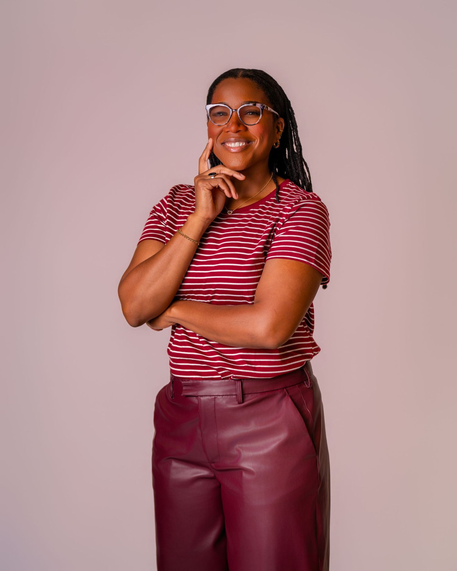 Smiling woman in striped red top and burgundy leather pants posing confidently with hand on chin.