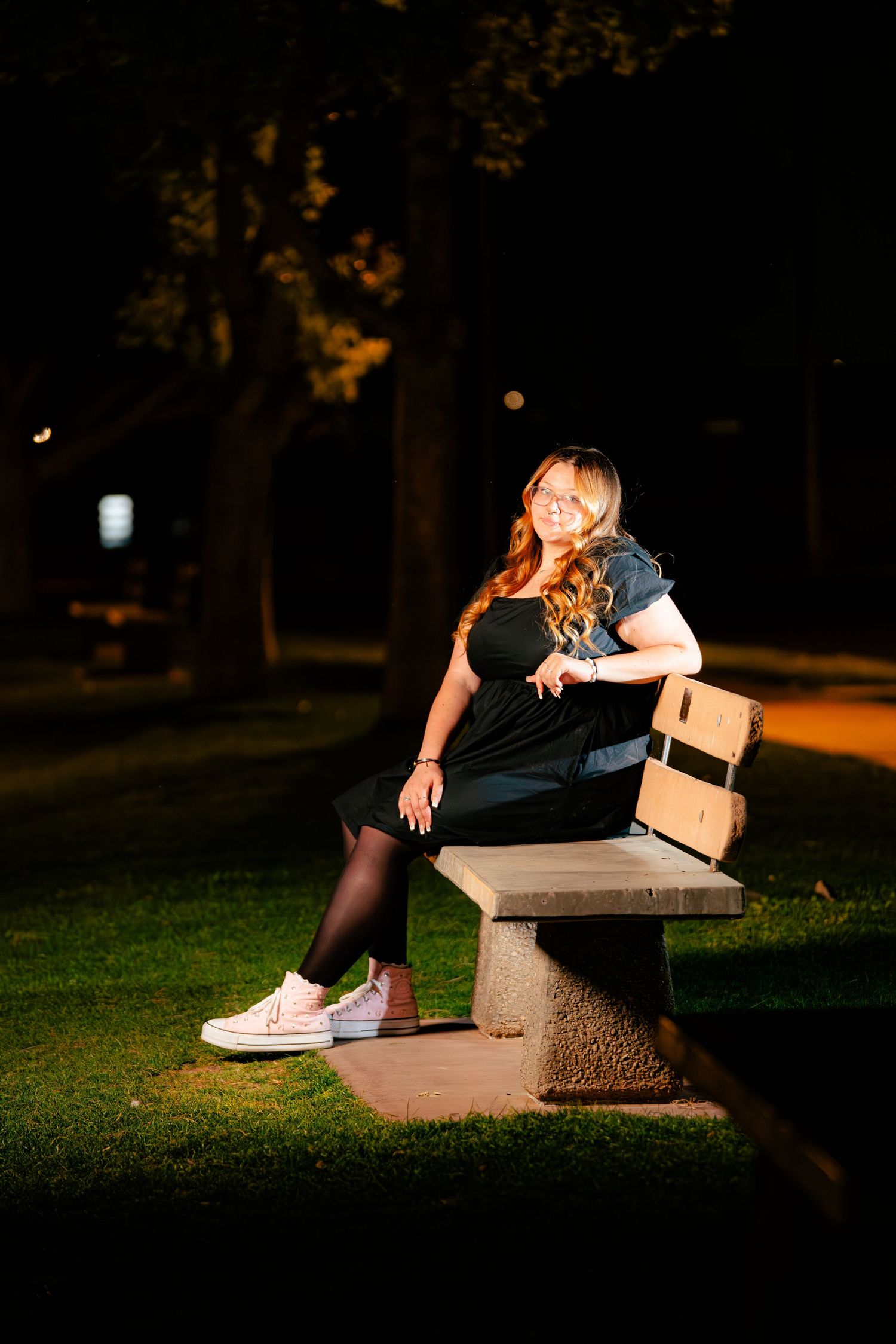 Young woman with red hair in black dress and white sneakers sitting on a park bench at night.