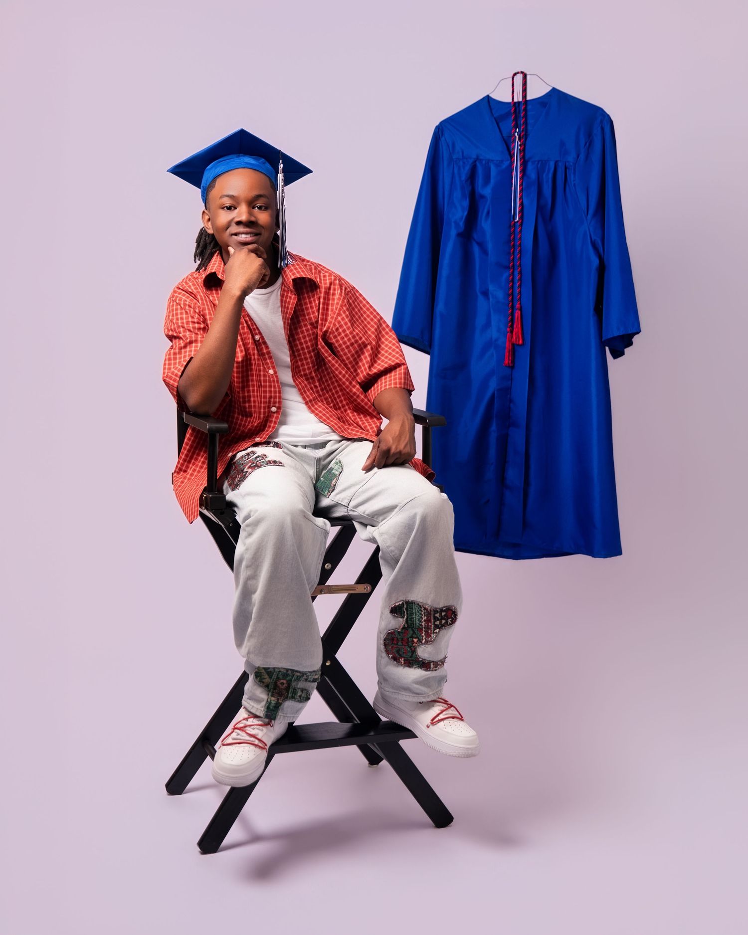 Young graduate wearing blue cap poses on chair beside hanging blue graduation gown with red tassel.