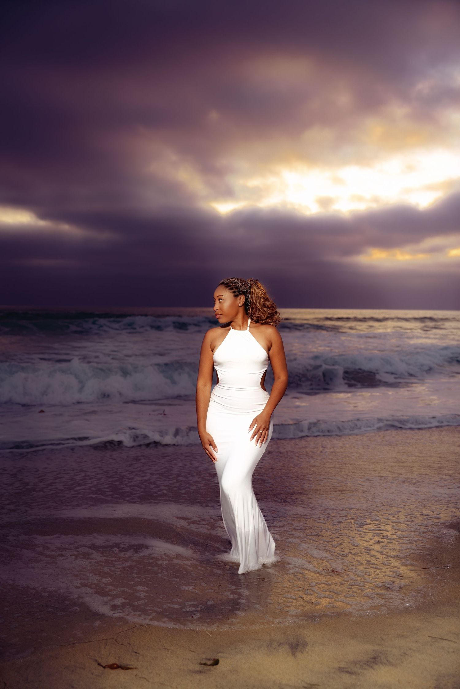 Dramatic beach photoshoot at sunset with flowing white dress in ocean waves.