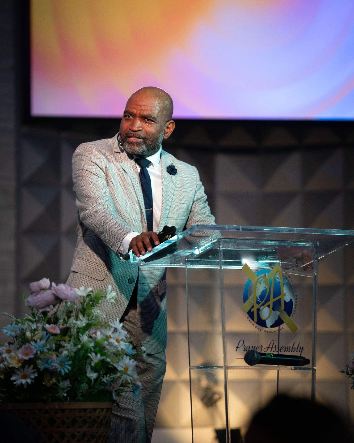 Speaker at clear glass podium delivers presentation with floral decorations on modern stage.